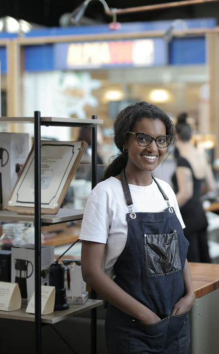 A barista wearing glasses and a dark apron stands behind a counter in a coffee shop, smiling at the camera.