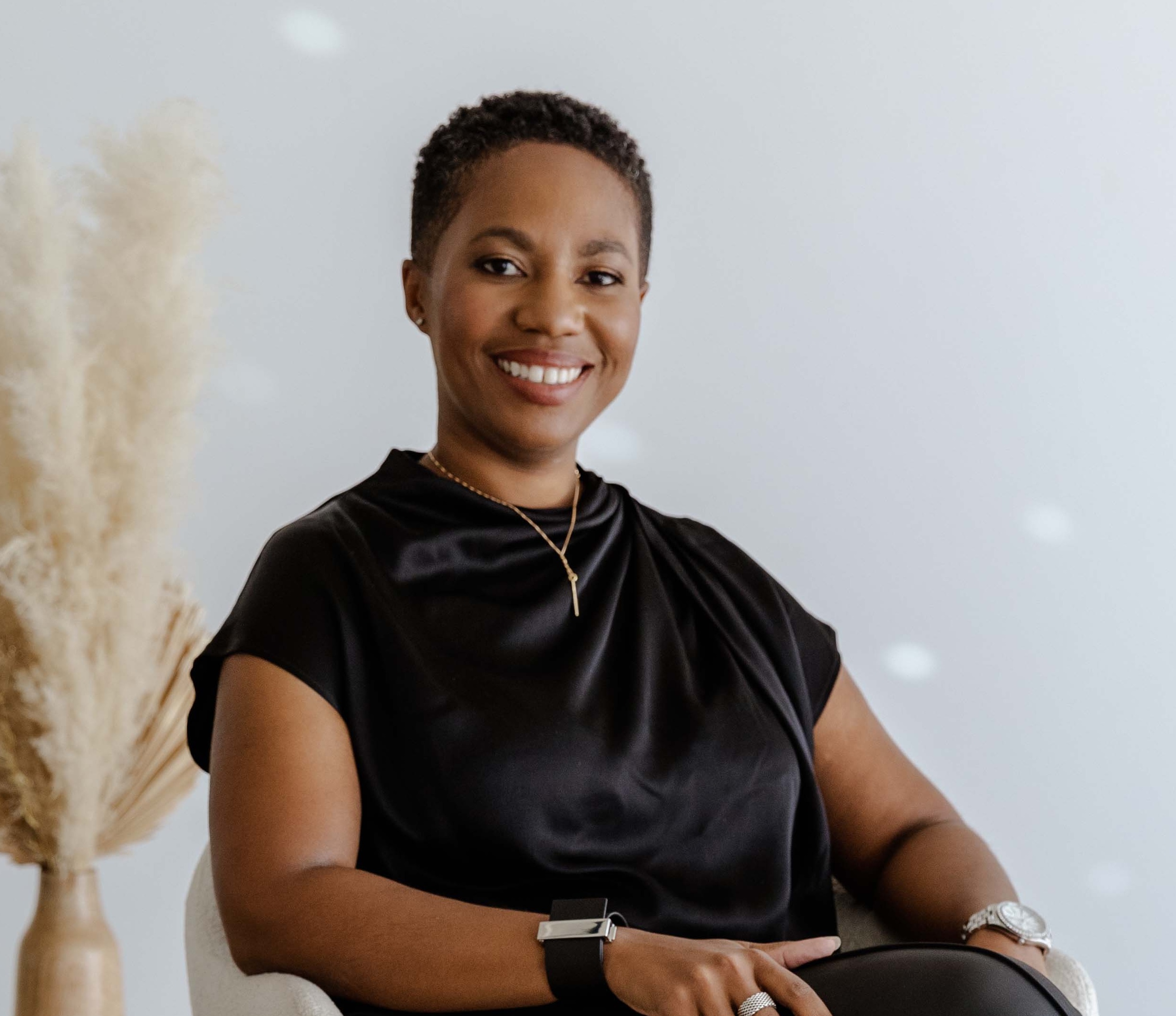 A person with short hair, wearing a black top and jewelry, sits and smiles at the camera. There is a vase with dried pampas grass in the background.