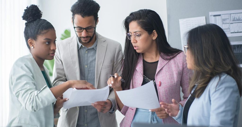 Four people standing and discussing documents in an office setting, with charts and graphs visible in the background.
