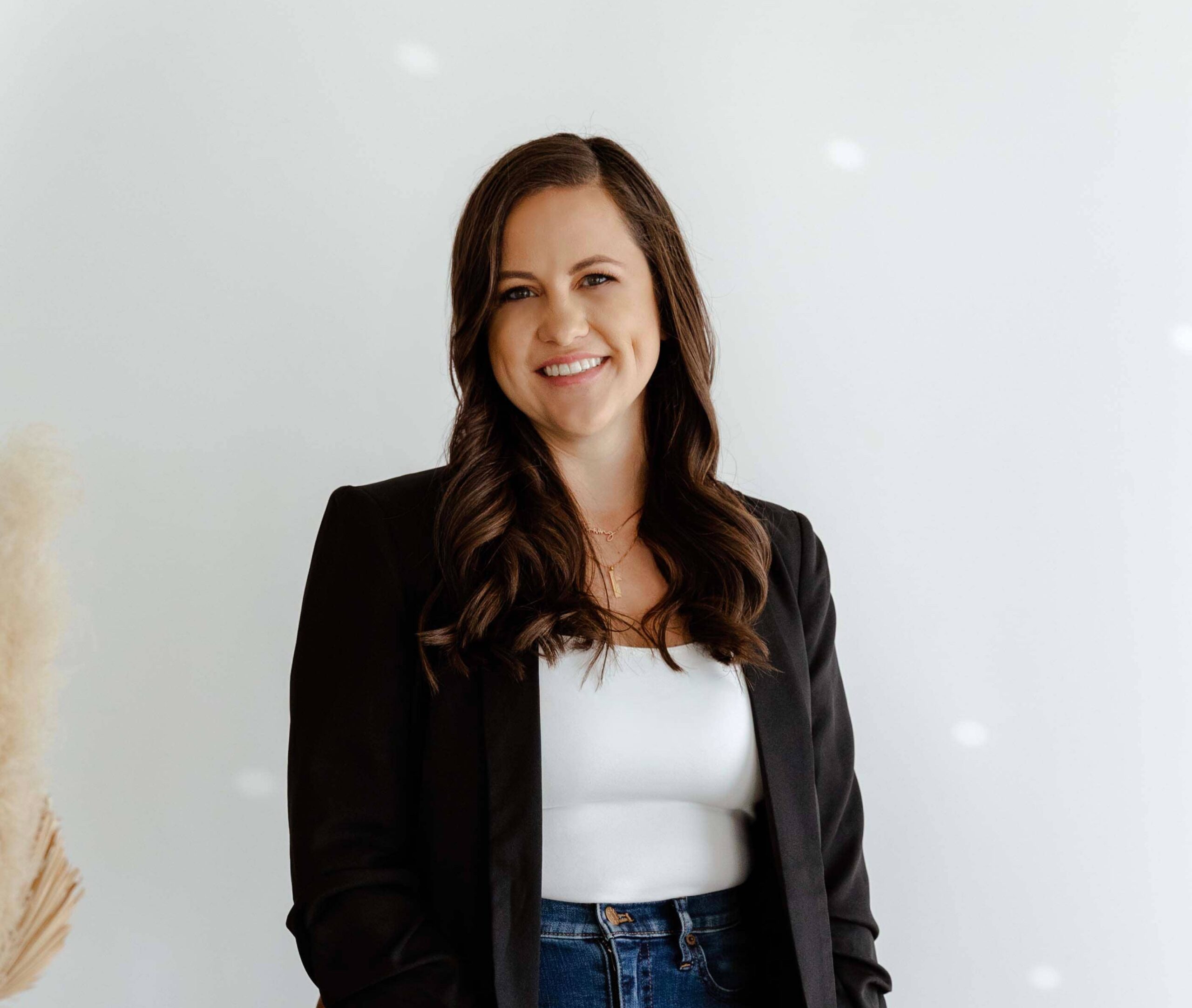 Woman with long brown hair wearing a black blazer, white top, and jeans, standing and smiling in front of a plain white background.