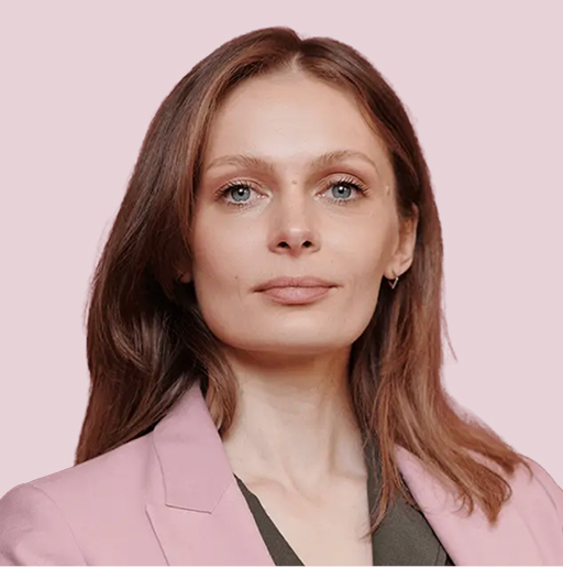 A woman with straight light brown hair, wearing a pink blazer over a dark top, poses against a solid light pink background.