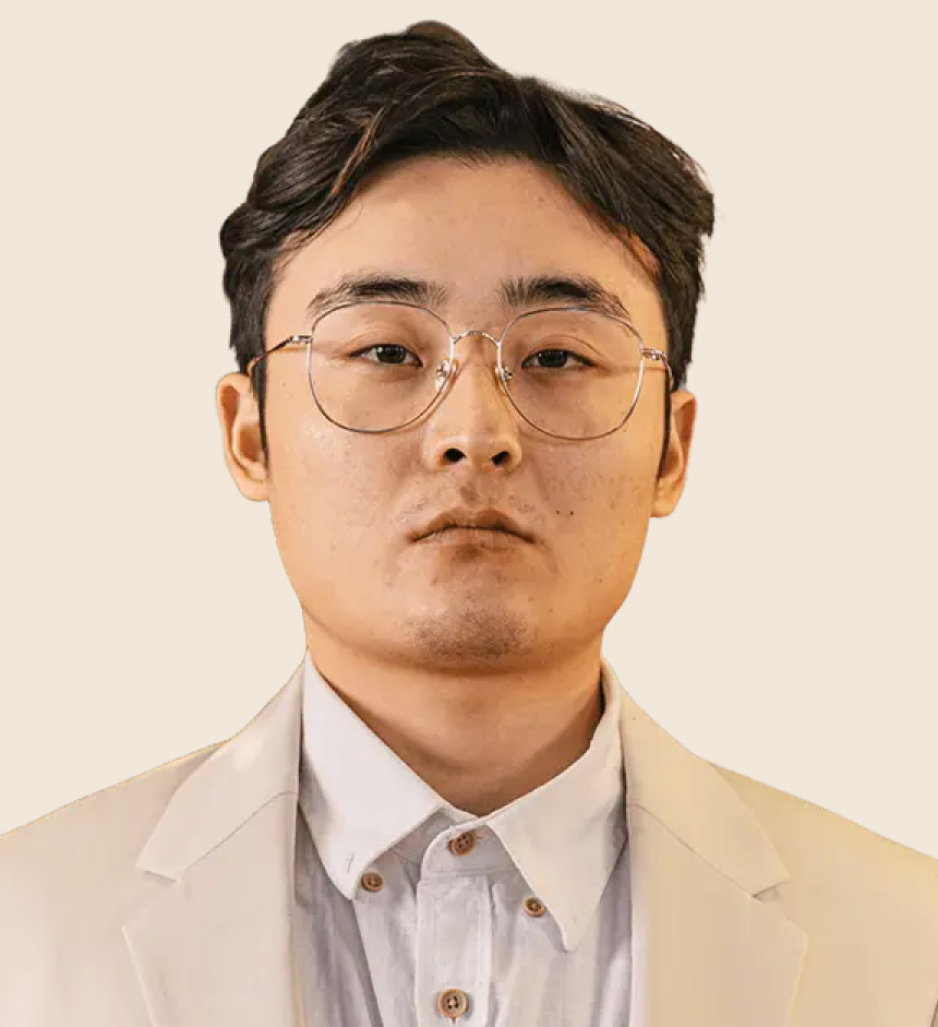 A young man with short dark hair and glasses wears a cream-colored suit and white shirt, facing the camera with a neutral expression against a plain background.