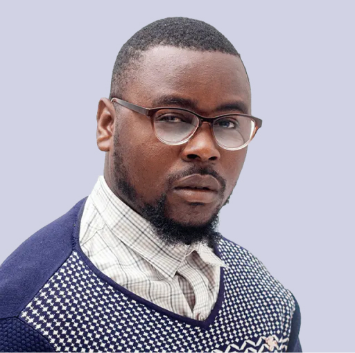 Man with short hair, beard, and glasses wearing a patterned sweater over a collared shirt, looking directly at the camera against a plain light background.