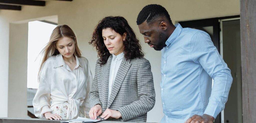 Three people stand outside by a railing, looking at documents together; one person appears to be explaining or pointing at the papers.