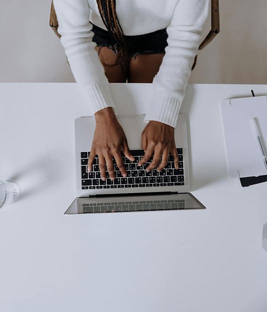 Person in a white sweater typing on a laptop at a white desk with a glass of water, clipboard, and pen nearby.