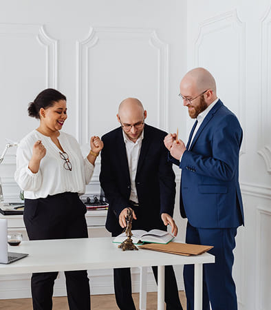 Three people in business attire stand around a desk in an office, appearing to celebrate or discuss something enthusiastically with open books and documents on the table.
