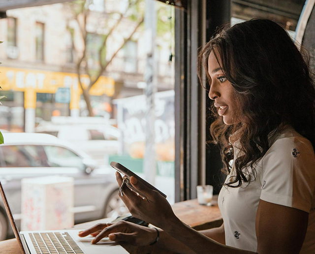 A woman sits by a window at a cafe, using a laptop and holding a smartphone, with city street and cars visible outside.